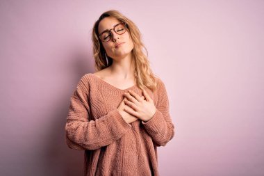 Genç, güzel sarışın kadın pembe arka planda gözlük ve süveter takıyor. Elleri göğsünde, gözleri kapalı gülümsüyor ve yüzünde minnet ifadesi var. Sağlık kavramı.