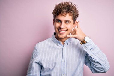 Sarışın, kıvırcık saçlı, çizgili tişörtlü, beyaz arka planda gülümseyen, el ve parmaklarıyla telefonla konuşan genç, yakışıklı bir adam. İletişim kavramları.