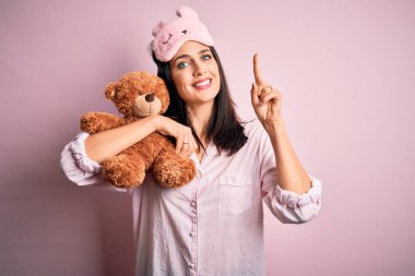 Mavi gözlü, pijama giyen, oyuncak ayıya sarılan genç esmer bir kadın bir fikir ya da soruyla şaşırmış, mutlu yüzlü, bir numara