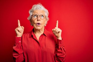 Son sınıf güzeli, gri saçlı, rahat gömlekli ve gözlüklü bir kadın kırmızı arka planda şaşkın şaşkın bakıyor, parmaklarıyla işaret ediyor ve kollarını kaldırıyordu..