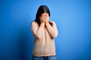 Mavi arka plan üzerine sıradan süveter giyen genç ve güzel bir kadın. Ağlarken suratını ellerle kapatan üzgün bir ifade. Depresyon kavramı.