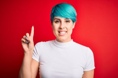 Mavi saçlı, kırmızı arka planda sıradan tişörtler giyen genç ve güzel bir kadın bir numaralı parmağını gösterirken kendinden emin ve mutlu bir şekilde gülümsüyor..