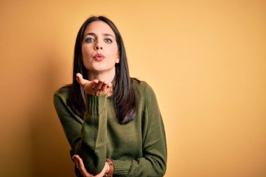 Sarı arka planda yeşil süveter giyen, mavi gözlü genç esmer kadın havadaki elle öpüşen kameraya bakıyor. Güzel ve seksi. Aşk ifadesi.