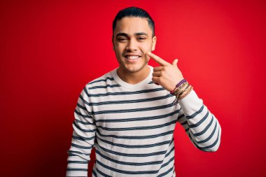 İzole edilmiş kırmızı arka planda dikilen sıradan çizgili tişört giyen genç Brezilyalı bir adam. El ve burunla işaret ediyor. Neşeli gülümsüyor. Güzellik konsepti