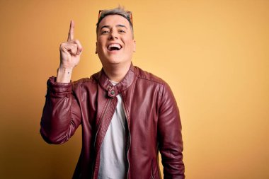 Sarı arka planda, deri ceket ve güneş gözlüğü takan yakışıklı, modern bir adam başarılı bir fikri işaret ediyor. Heyecanlı ve mutlu. Bir numara..