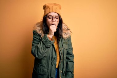 Gözlüklü esmer kadın ve sarı izole edilmiş arka planda şapkalı kışlık kadın kendini kötü hissediyor ve soğuk algınlığı veya bronşitin belirtisi olarak öksürüyor. Sağlık hizmeti kavramı.