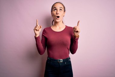 İzole edilmiş pembe arka plan üzerinde duran gündelik tişört giyen genç ve güzel bir kadın şaşırmış ve şaşırmış bir şekilde yukarı bakıyor ve parmaklarıyla işaret ediyor ve kollarını kaldırıyordu..