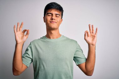 İzole edilmiş beyaz arka plan üzerinde duran sıradan tişört giyen genç yakışıklı adam rahatla ve parmaklarıyla meditasyon hareketi yaparken gözleri kapalı gülümse. Yoga konsepti.