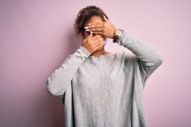 Genç ve güzel Afro-Amerikalı kız pembe arka planda süveter ve gözlük takıyor gözleri ve ağzı kapalı, şaşırmış ve şok olmuş. Duygularını gizliyor.