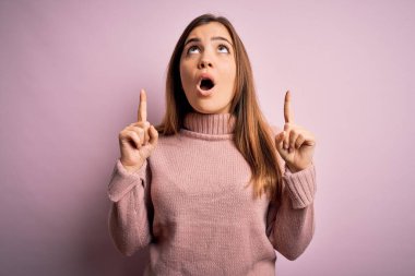 Güzel genç bir kadın pembe izole edilmiş arka planda balıkçı yaka kazak giyiyor. Şaşırmış ve şaşırmış bir şekilde yukarı bakıyor ve parmaklarıyla işaret ediyor ve kollarını kaldırıyor..