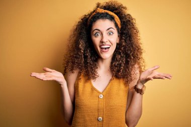 Kıvırcık saçlı, piercing takan, gözlüklü, kolları çapraz gülümseyen ve kameraya bakan güzel esmer kadın. Pozitif kişi.