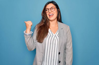 Genç İspanyol iş kadını mavi izole edilmiş arka planda duran gözlüklü mutlu yüzüyle gülümseyen ve başparmağıyla yan tarafı gösteren..