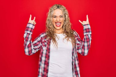Genç, sarışın, sıradan bir gömlek giyen, izole edilmiş kırmızı arka planda duran çılgın bir ifadeyle el kaldırarak rock sembolü yapan genç bir kadın. Müzik yıldızı. Ağır müzik konsepti.