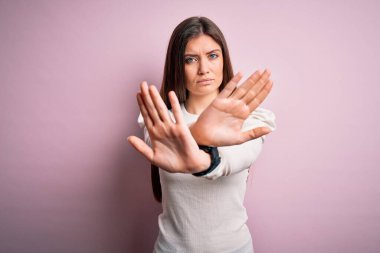 Mavi gözlü genç bir kadın pembe arka planda günlük beyaz tişört giyiyor. Reddedilmiş yüz ifadesi, kollar ve avuç içleri kesişmiş, kızgın yüz ifadesi takınıyor.