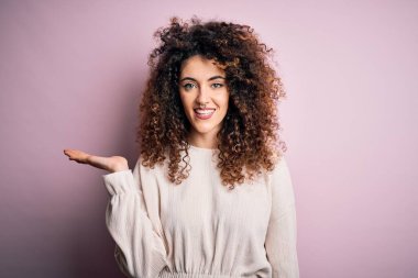 Kıvırcık saçlı ve piercingli güzel bir kadın pembe arka planda rahat bir süveter giyiyor neşeli bir şekilde gülümsüyor ve avuç içi kameraya bakıyor..