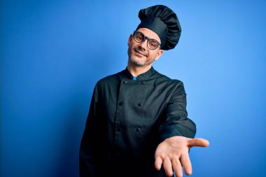 Genç, yakışıklı aşçı üniformalı ve şapkalı, mavi arka planda neşeli gülümsemeli, yardım eli uzatan ve kabul gören bir aşçı..