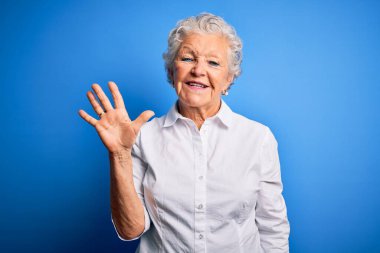 Üst düzey güzel bir kadın zarif bir gömlek giyer mavi arka plan üzerinde durur ve parmaklarıyla beş numarayı gösterirken kendinden emin ve mutlu bir şekilde gülümser..