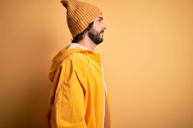 Sakallı genç yakışıklı adam yağmurluk giyiyor sarı arka planda yağmurluk giymiş, rahat bir görünüm takınıyor, kendine güvenen bir gülümsemeyle doğal bir poz veriyor..