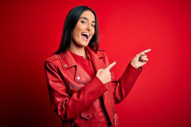 Genç, güzel, esmer, sıradan bir ceket giyen, izole edilmiş kırmızı arka planda duran, gülümseyen ve kameraya bakan iki eliyle ve parmaklarıyla yüzünü gösteren bir kadın..