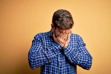 Sarışın, sakallı ve mavi gözlü genç bir işadamı sarı arka planda gömlek giyiyor ve ağlarken suratını ellerle kapatıyor. Depresyon kavramı.