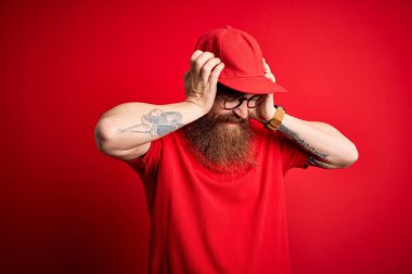 Gözlük takan yakışıklı bir teslimatçı ve izole edilmiş arka planda kırmızı şapkalı. Ağrı ve migrenden dolayı çaresiz ve stresli. Ellerini başının üstüne koy..