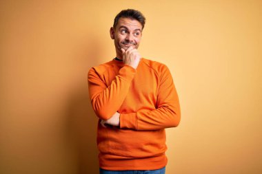 Turuncu günlük süveter giyen genç yakışıklı adam, izole edilmiş sarı arka planda duruyor, elleri çenesinde soru sorarken, düşünceli bir ifade takınıyor. Düşünceli bir yüzle gülümsüyor. Şüphe kavramı.