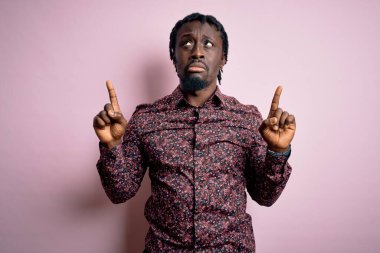 Genç, yakışıklı, Afro-Amerikalı bir adam pembe arka plan üzerinde dikilmiş üzgün ve üzgün görünüyor, parmaklarıyla yön gösteriyor, mutsuz ve depresif..