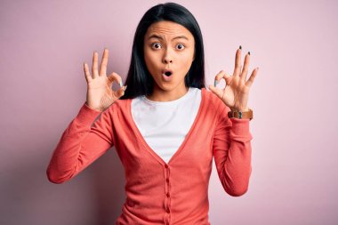 İzole edilmiş pembe arka plan üzerine sıradan süveter giyen genç ve güzel bir Çinli kadın parmaklarıyla onay sembolü yaparken şaşırmış ve şok olmuş görünüyor. Çılgın ifade