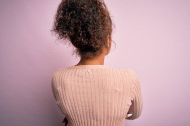 Genç, güzel, Afro-Amerikalı bir kız pembe arka planda dikilip kollarını kavuşturup arkasına bakıyor.