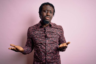 Genç, yakışıklı, Afro-Amerikalı bir adam pembe arka planda dikilmiş, habersiz ve kolları açık, hiçbir fikri yok..