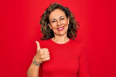 Orta yaşlı, esmer, sıradan tişörtlü bir kadın kırmızı arka planda dikiliyor ve elleriyle mutlu bir jest yapıyor. Kameraya bakıp başarıyı gösteren ifade onaylanıyor.