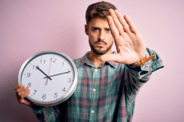 Sakallı genç adam geri sayım yapıyor. Tecrit edilmiş pembe arka plan üzerinde büyük bir saat kullanıyor. Açık elle dur işareti yapıyor.