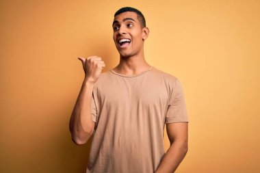 Genç, yakışıklı, Afro-Amerikalı, günlük tişörtler giyen, sarı arka planda dikilen mutlu yüzüyle gülümseyen ve baş parmağıyla yüzünü gösteren bir adam..