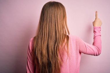 Genç, güzel, kızıl saçlı bir kadın, pembe arka planda rahat bir süveter giyiyor. Parmak uçlarıyla geriye doğru poz veriyor.