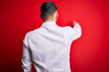 Mavi gözlü, zarif gömlekli genç bir iş adamı kırmızı izole edilmiş arka planda dikiliyor ve parmağıyla öne doğru poz veriyor.