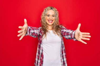 Genç, sarışın, gündelik bir gömlek giyen, izole edilmiş kırmızı arka planda duran ve kucaklaşmak için kollarını açıp gülümseyen bir kadın. Mutluluğu kucaklayan neşeli bir ifade..