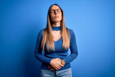 Mavi gözlüklü, genç, sarışın bir kadın. Mavi arka planda duruyor. Midesi bulanıyor, midesi bulanıyor. Ache kavramı.