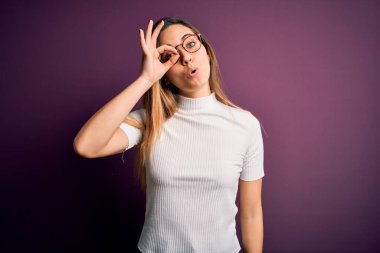 Mor arka planda sıradan tişörtler giyen, mavi gözlü genç, güzel sarışın kadın şaşırmış yüz ifadesiyle şaşırmış, parmaklarından bakan bir jest yapıyor. İnançsız ifade.