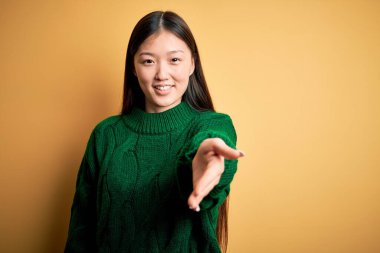 Sarı arka planda yeşil kış süveteri giyen güzel Asyalı bir kadın neşeli bir gülümsemeyle yardım eli uzatıyor ve kabul ediyor..