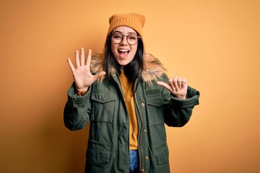 Genç esmer kadın, gözlük takıyor ve şapkalı, sarı izole edilmiş arka planı var. Altıncı parmaklarını göstererek gülümsüyor. Kendinden emin ve mutlu..