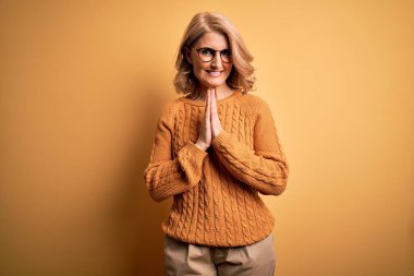 Orta yaşlı güzel sarışın kadın sarı arka planda günlük süveter ve gözlük takıyor birlikte dua edip af diliyor kendinden emin bir şekilde gülümsüyor..