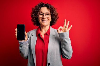 Orta yaşlı kıvırcık kadın elinde akıllı telefonuyla izole edilmiş kırmızı arka planda ekranı gösteriyor. Tamam işareti yapıyor, mükemmel bir sembol.