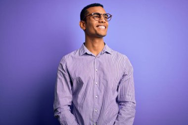 Yakışıklı Afro-Amerikalı adam mor arka plan üzerinde çizgili gömlek ve gözlük takıyor. Yüzünde gülümseme ve doğal ifadeyle diğer tarafa bakıyor. Kendinden emin gülen.