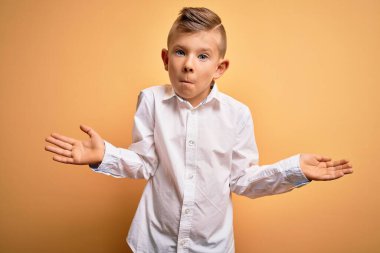 Küçük, beyaz, mavi gözlü, sarı arka planda zarif beyaz gömlek giyen kolları ve elleri havada, kafası karışmış bir çocuk. Şüphe kavramı.