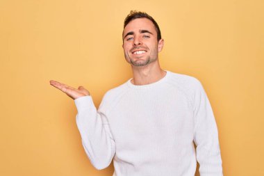 Mavi gözlü, sıradan bir süveter giyen, sarı arka planda durup gülümseyen, elinde avucuyla kameraya bakan genç yakışıklı bir adam..