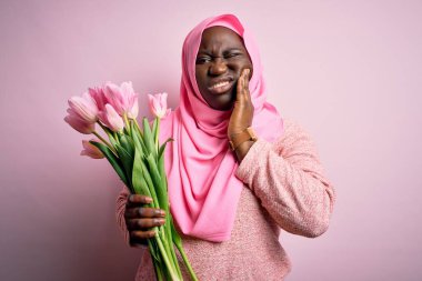 Genç Afro-Amerikan artı cüsseli kadın diş ağrısı ya da diş ağrısı yüzünden pembe lalelerle dolu buketi tutup ağızlarına dokunuyor. Dişçi konsepti.