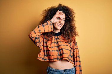 Kıvırcık saçlı ve piercingli genç ve güzel bir kadın. Günlük tişört giyiyor. El hareketleriyle gülümsüyor..