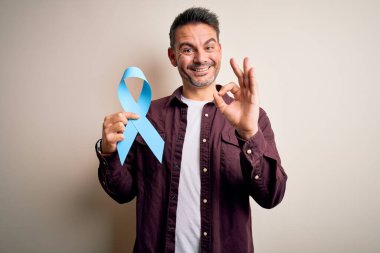 İzole edilmiş beyaz arka planda mavi kanser kurdelesi tutan genç yakışıklı adam tamam işareti yapıyor, mükemmel bir sembol.