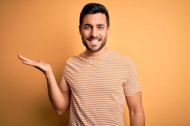 Sarı arka planda sıradan çizgili tişörtler giyen sakallı genç yakışıklı bir adam gülümseyen, eli kameraya bakan ve elini gösteren..