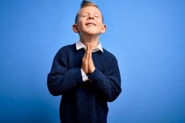 Mavi arka planda mavi süveter giyen, elleri bağlı yalvaran ve dua eden, yüzünde umut ifadesi olan, mavi gözlü küçük beyaz çocuk çok duygusal ve endişeli. Yalvarıyorum..